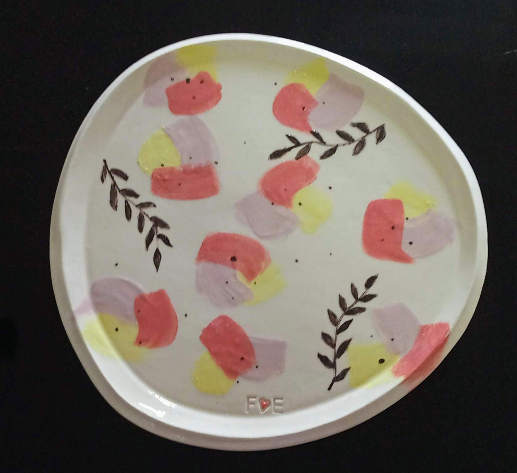 Plato mediano de forma irregular, de fondo color hueso y decoración con engobe negro en forma de hojas pequeñas y manchas de colores amarillo, lila y coral pastel